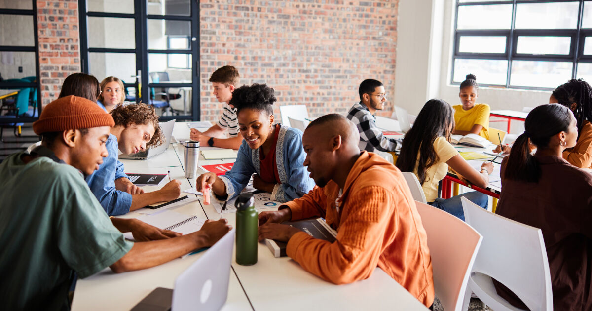 A group of teacher candidates sit at desks during a graduate class and talk with one another. Some of the students take notes.